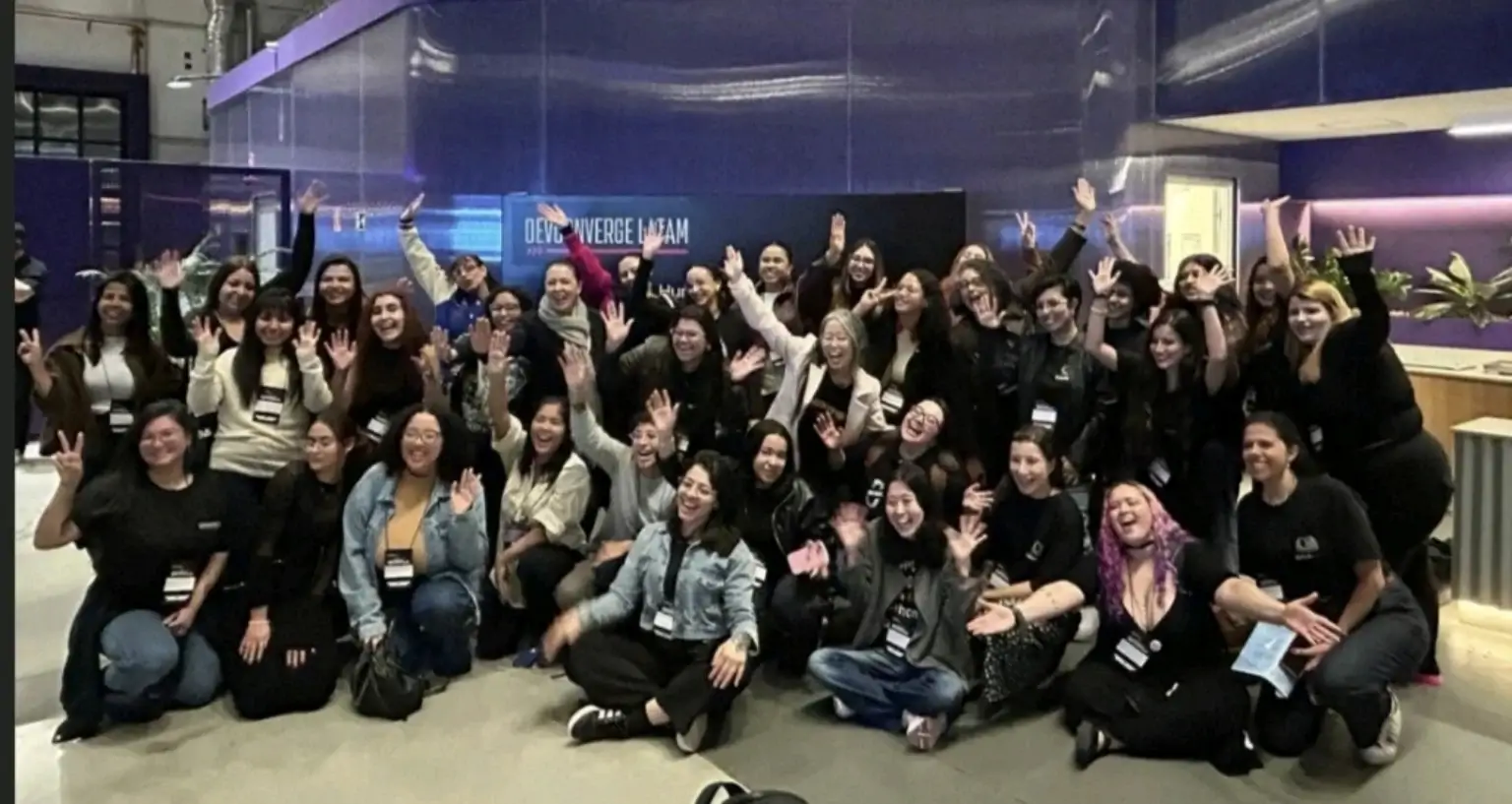 Grupo de participantes, em sua maioria mulheres, acenando para a câmera num evento de tecnologia